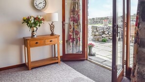 Una habitación con una consola de madera, un reloj de pared, una puerta con cortina floral y vistas a una pared de piedra y follaje exterior.