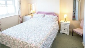 Quarto bem arrumado com colcha florida, mesa de cabeceira com abajur, cadeira e janela com cortinas.