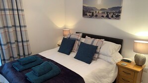 A neatly made bed with a dark patterned bedspread, multiple pillows, and folded towels on a headboard. There is a bedside table with a lamp and a remote control. A painting of a mountainous landscape hangs on the wall above the bed. - 2 bedroom accommodation in Keswick (Keswick)