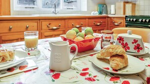 Une cuisine avec des meubles en bois, une fenêtre donnant sur un beau paysage, une table recouverte d’une nappe à motifs floraux et un saladier rempli de fruits.