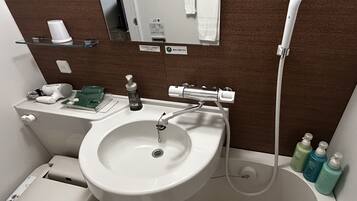 A bathroom with a sink, mirror, and shower. There are toiletries on the sink and a hairdryer on the floor.