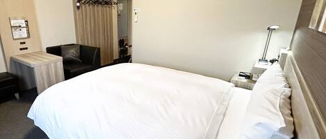 A hotel room with a large bed, a chair, a small table, and a nightstand with a lamp.