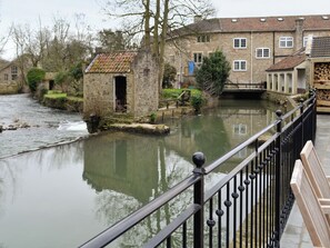 A stone bridge over a calm river, with stone buildings and a small wooden structure on the riverbank.