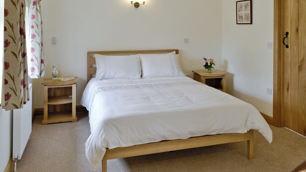 A neatly made bed with white linens, a bedside table with a vase of flowers, and a wall-mounted light fixture. - 3 bedroom accommodation in Spring Gardens, Frome (Spring Gardens, Frome)