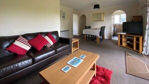 A living room with a leather sofa, a wooden coffee table, a dining table with chairs, a television on a wooden stand, and a red rug. - 3 bedroom accommodation in Liskeard (Liskeard)