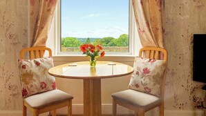 A room with two wooden chairs, a small round table with a vase of flowers, and a window with curtains. - Old Barn Farm Cottage - CC539048 (Uplawmoor, near Paisley)