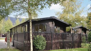A wooden cabin with a deck, surrounded by trees and a grassy area. - Ewe How - Burnside Park (Keswick)