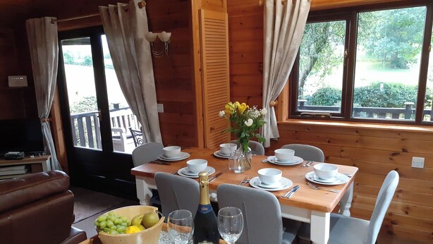 A dining area with a wooden table set for four, a bowl of fruit, a bottle of champagne, and a vase with flowers. - Ewe How - Burnside Park (Keswick)