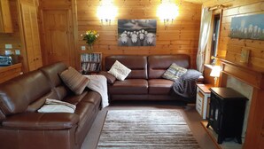 A cozy living room with a brown leather sofa, a coffee table, and a fireplace. There is a bookshelf with books and a vase with flowers. - Ewe How - Burnside Park (Keswick)