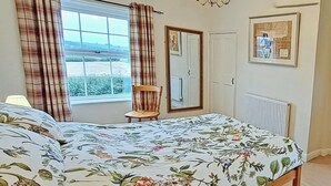 A bedroom with a floral bedspread, a wooden chair, a window with curtains, a radiator, and a framed picture on the wall. - Foston Grange Cottage (Foston near York)