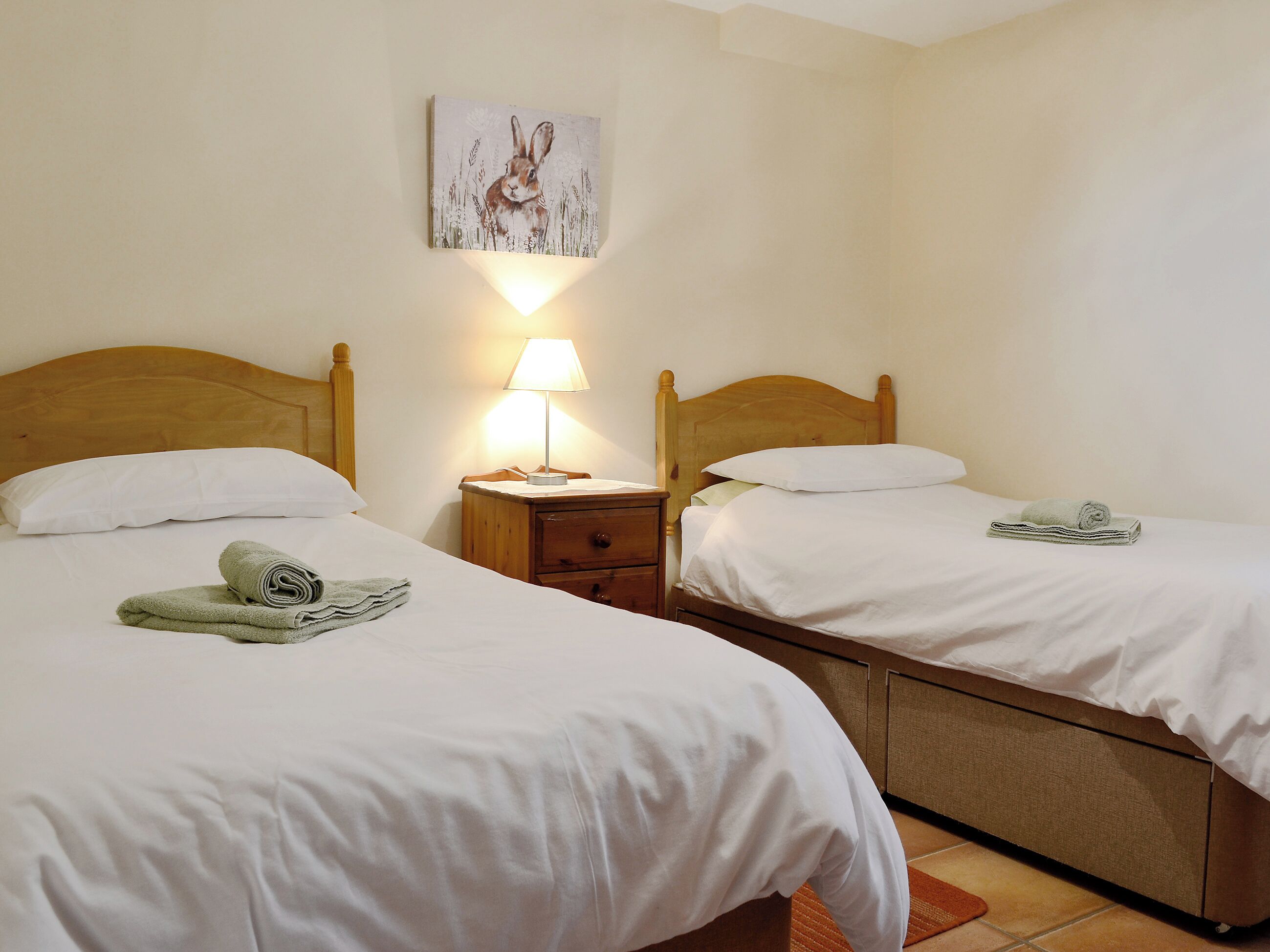 A hotel room with two beds, a nightstand with a lamp, a painting on the wall, and a rolled towel on each bed.