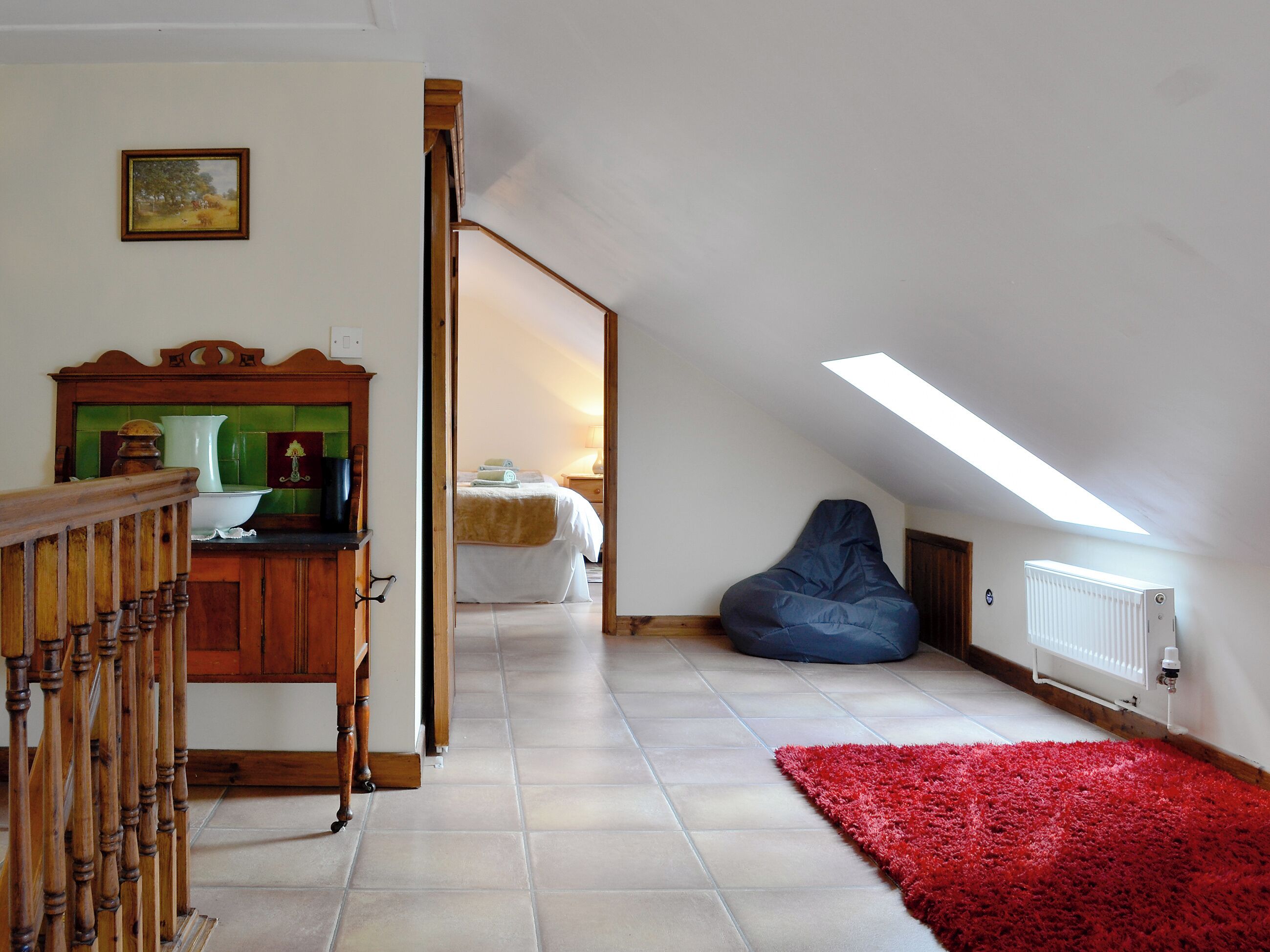 Una stanza con soffitto spiovente, un letto, un tappeto rosso, un pouf blu e un armadietto in legno.
