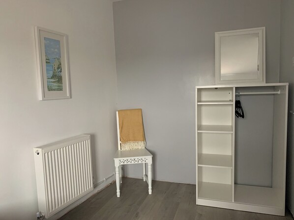 A room with a white radiator, a small white chair with a brown cushion, and a white wardrobe with shelves. - 2 bedroom accommodation in Haverigg, near Millom (Haverigg, near Millom)