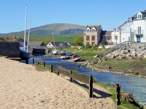 A riverside scene with boats docked, a sandy beach, and buildings with a hill in the background. - 2 bedroom accommodation in Haverigg, near Millom (Haverigg, near Millom)