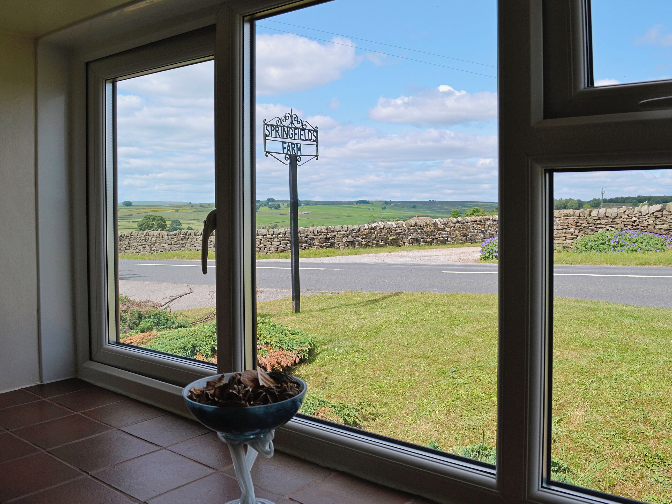 A view of a rural landscape with a stone wall and a road, seen through a window with a bowl of dried flowers on a stand.