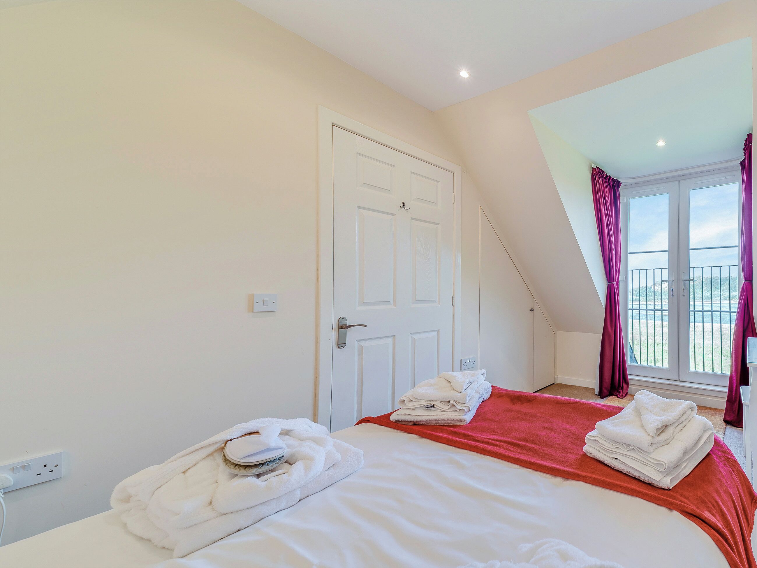 A bedroom with a bed, a window with curtains, a door, and a power outlet.