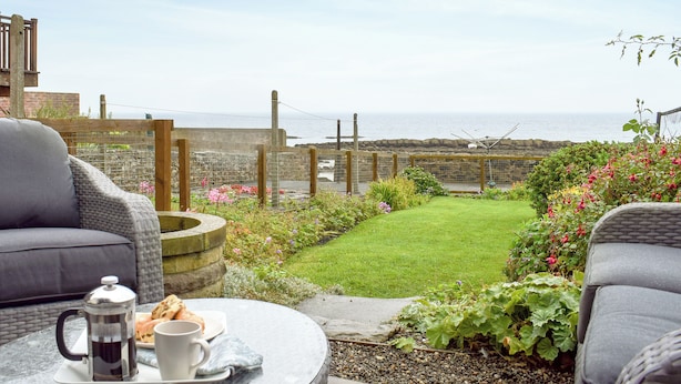 Une terrasse avec vue sur la mer, une table en pierre avec une cafetière et des pâtisseries, et des meubles en osier.