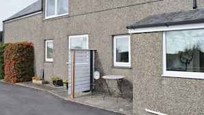 A two-story house with a gravel exterior, multiple windows, and a small patio area with a table and chair. - Fron Erch Cottages - 2712 (Abererch, near Pwllheli)