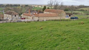 Un bâtiment en pierre comprenant plusieurs pièces, entouré d’une zone délimitée par une clôture et d’un champ herbeux.