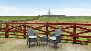 Two wicker chairs with cushions at a wooden fence overlooking a green field.