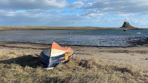 Una playa con un bote en la orilla, una estructura distante que parece un castillo y un mar tranquilo.