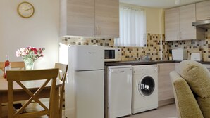 A compact kitchen with a refrigerator, microwave, washing machine, and a dining area with a chair and table. - Sally's Nest - E3042 (Wenhaston, near Southwold)