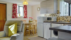 A compact kitchen with a refrigerator, microwave, and dishwasher. There is a dining table with chairs, a sofa with a cushion, and a window with curtains. - Sally's Nest - E3042 (Wenhaston, near Southwold)