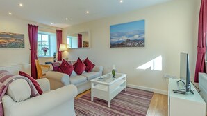 A living room with a white sofa, red cushions, a coffee table, and a painting of a snowy landscape. - Kestrel Cottage - 27937 (Stirling)