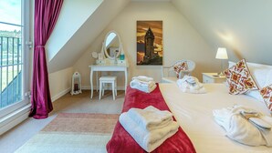 A bedroom with a bed, a desk with a mirror, a chair, a nightstand with a lamp, and a painting on the wall. - Kestrel Cottage - 27937 (Stirling)