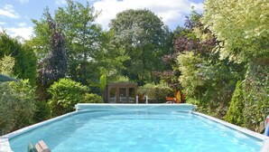 Una piscina con bordo in legno, circondata da vegetazione rigogliosa e un'area salotto coperta.