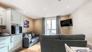 Un espacio habitable compacto con kitchenette, un sofá gris, una mesa de comedor y un televisor montado en la pared.