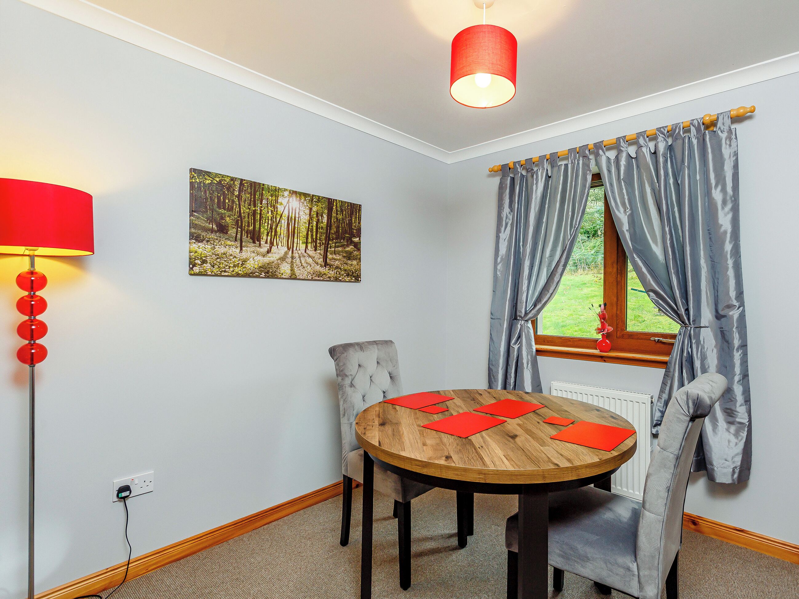 A dining area with a round wooden table, two grey upholstered chairs, a red floor lamp, a forest-themed wall art, and a window with grey curtains.