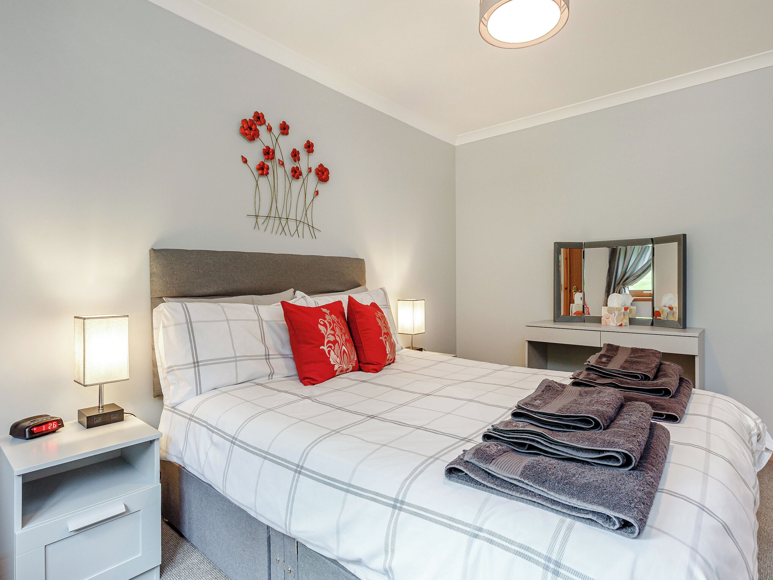 Una cama bien hecha con una colcha escocesa, dos almohadas rojas y una pila de toallas dobladas. Hay una mesita de noche con una lámpara y un reloj digital. Se ve una obra de arte en la pared con flores rojas sobre la cama.