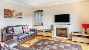 A modern living room with a leather sofa, a fireplace, a flat-screen TV, and a large window with blinds. - 1 bedroom accommodation in Achaphubuil near Fort William (Achaphubuil near Fort William)