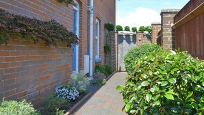 Une ruelle étroite pavée de briques, avec un chemin, une clôture en bois et diverses plantes et fleurs.