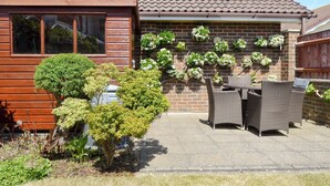 Une maison d’habitation avec un toit en tuiles, un mur en briques et un patio aménagé avec des meubles en osier et des plantes en pot.