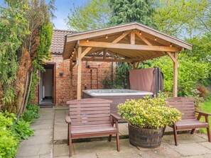 Una pérgola de madera con luces colgantes, un jacuzzi y dos bancos de madera en un jardín.