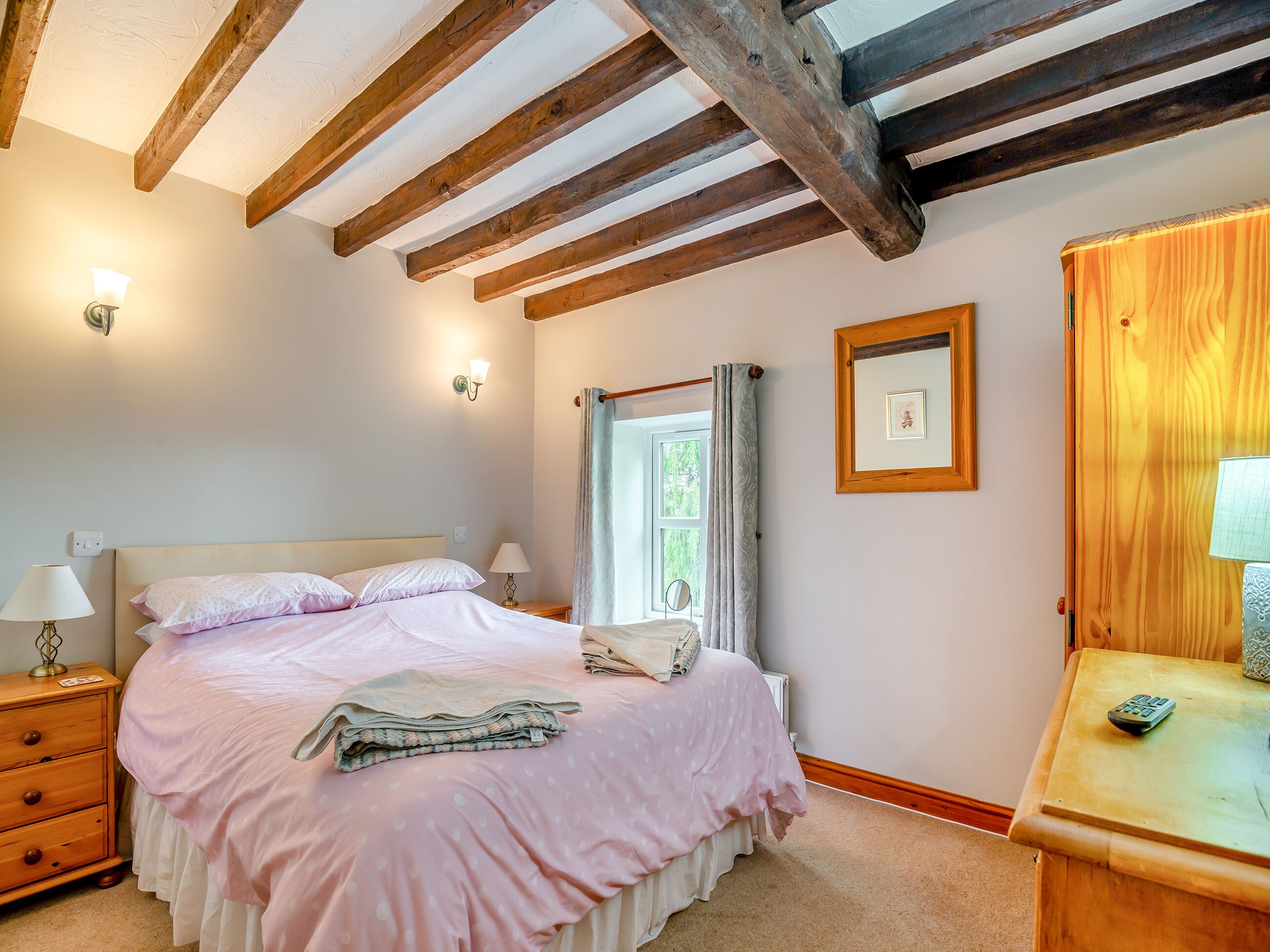 A bedroom with a bed, bedside lamps, a wooden headboard, a window with curtains, a framed picture, and a wooden nightstand with a lamp.