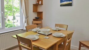 A dining area with a wooden table set for four, surrounded by chairs. A window with curtains is visible, and a painting hangs on the wall.