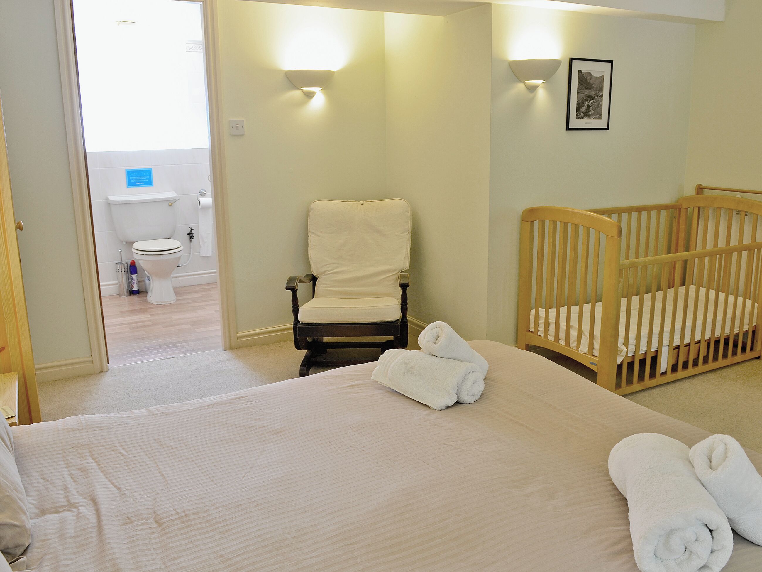 A bedroom with a bed, a wooden crib, a chair, and a toilet visible in the adjacent bathroom.
