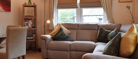 Una sala de estar con sofá, sillones, una mesa de centro de vidrio, estantería y ventana con cortinas.