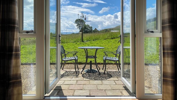 Une pièce dotée d’une porte vitrée donnant sur un patio extérieur avec une table et des chaises, et offrant une vue sur un champ verdoyant et un arbre.