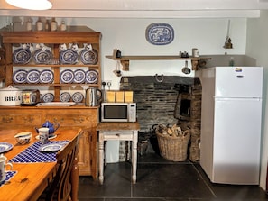 A kitchen with a microwave, toaster, kettle, and a refrigerator. There is a wooden table with blue and white plates and a polka dot placemat. A stone wall and a hanging light fixture are also visible.