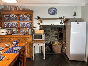 La cuisine est équipée d’un four à micro-ondes, d’un grille-pain, d’une bouilloire et d’un réfrigérateur. On y trouve une table en bois avec des assiettes bleues et blanches, ainsi qu’une nappe à pois. Un mur en pierre et un luminaire suspendu sont également visibles.