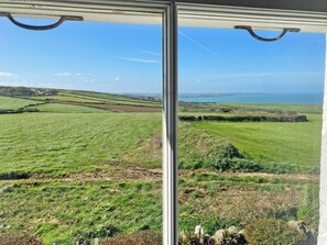 Blick durch ein Fenster auf eine ländliche Landschaft mit Feldern, einem entfernten Gewässer und einem klaren Himmel.