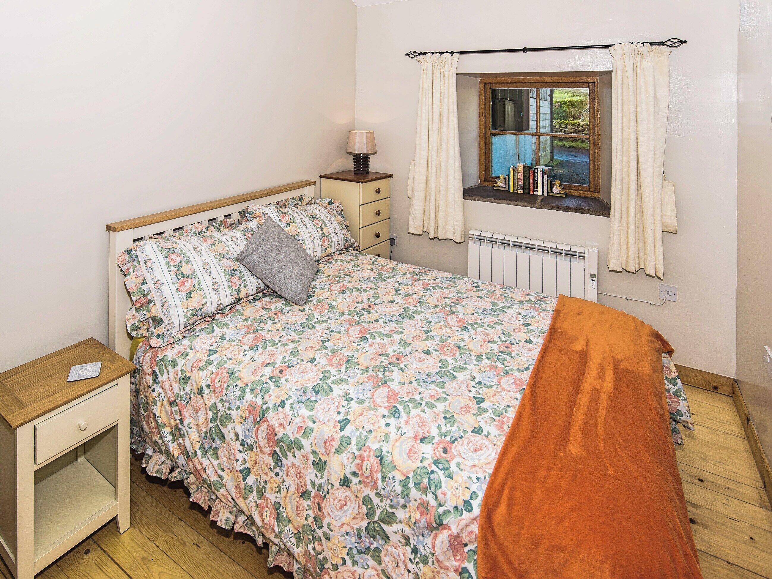 A bedroom with a floral bedspread, a wooden nightstand, a dresser, a window with curtains, and a radiator.