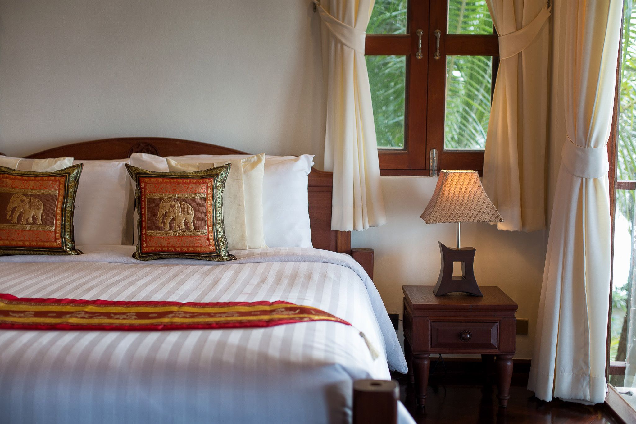 Una cama bien hecha con ropa de cama a rayas, dos almohadas decorativas, una lámpara de noche y vistas a la vegetación a través de una ventana.