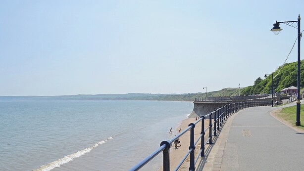 A seaside promenade with a paved walkway, a stone sea wall, and a metal railing.