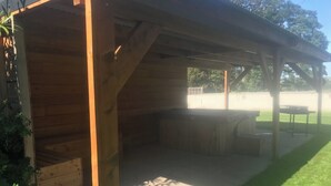 Un patio techado al aire libre con una parrilla y zona de estar, rodeado por una estructura de madera y un césped.