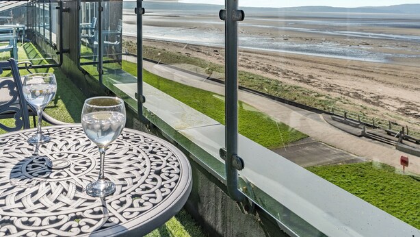 Une table avec un verre d’eau et un verre à vin, donnant sur une plage bordée d’une rambarde en métal.
